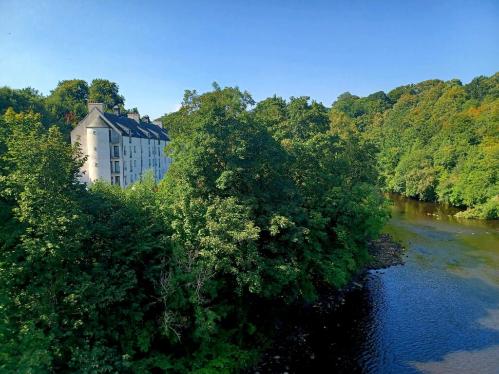 Old mill building and riverbank at the David Livingstone Centre, partially hidden among trees beside the river Clyde.